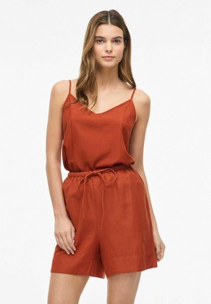 Kobieta ubrana w bezrękawnikowy, rdzawego koloru kombinezon z regulowanymi ramiączkami i sznurkiem w talii, stojąca na jednolitym białym tle.
