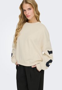 Femme blonde aux cheveux ondulés lâchés portant un sweat-shirt crème oversize avec des lettres noires sur les manches et un pantalon large noir, les mains dans les poches.