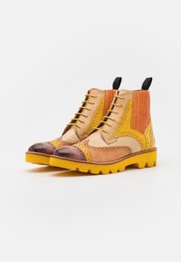Botines bicolor con cuero beige, naranja y amarillo, detalles de perforado, puntera marrón y suela amarilla gruesa.