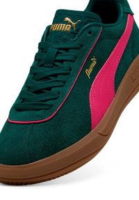 Grüne Wildledersneaker mit einem pinken seitlichen Streifen, goldenem Puma-Logo und brauner Gummisohle. Verfügt über runde Schnürsenkel und eine strukturierte Zunge.