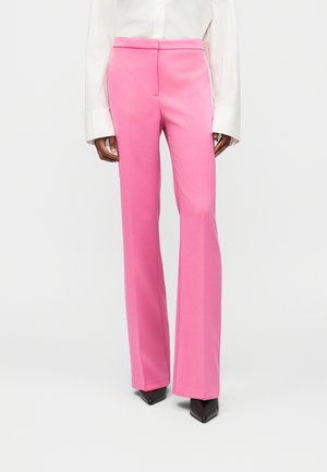 Naine, kes kannab kõrge vöökohaga, sirgete säärtega erkroosa pükse, valget pika varrukaga särki ja musti teravate ninadega kingi.