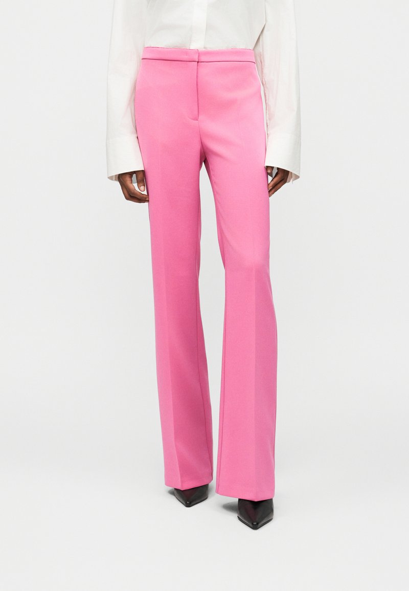 Γυναίκα που φοράει παντελόνι ψηλόμεσο, ίσιο με έντονο ροζ χρώμα, λευκό πουκάμισο μακριάς μανίκας και μαύρα μυτερά παπούτσια.