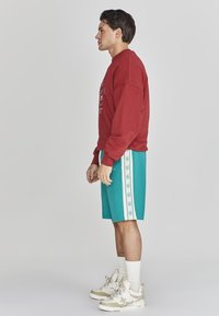 Felpa rossa con design grafico, abbinata a pantaloni corti turchesi con strisce bianche laterali e testo. Indossata con sneaker beige e calze bianche.