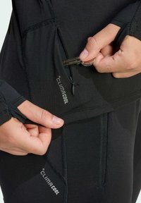 Sort atletisk top med tætsiddende design og tommelfingerhuller. Har en lynlåslomme til små genstande, mærket "climacool" nær lynlåsen.