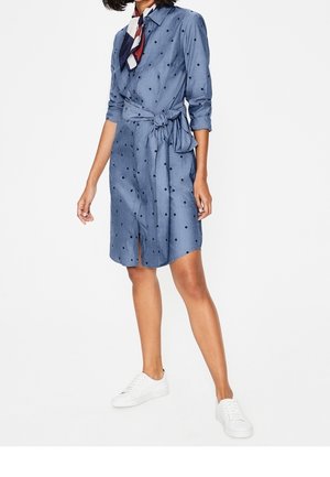 Woman wearing a blue polka dot shirt dress with a tie waist, a multicolored neck scarf, and white sneakers standing against a white background.