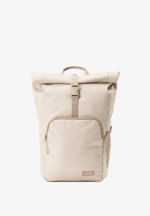 Sac à dos beige à rabat fabriqué en tissu durable, avec une poche zippée à l'avant, une sangle réglable et une poignée supérieure pour un transport facile.