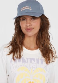 Blue cap with embroidered "O'Neill" logo in a bronze tone, featuring a structured design and a curved brim. Worn with a white graphic t-shirt.