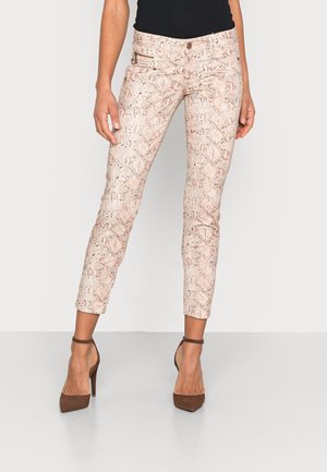 Woman wearing light pink snake print slim-fit ankle pants and brown high-heeled ankle strap shoes, standing against plain background.