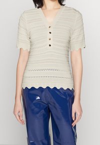 Béžový krátký rukáv s výstřihem do V, se zvlněným okrajem, strukturovanými pruhy a ozdobnými knoflíky vpředu. Nošený spolu s lesklými modrými kalhotami.