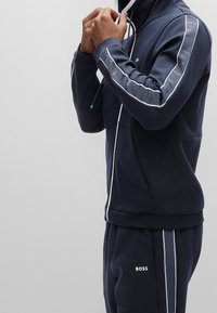 Navyblå tvådelad träningsoverall i mjukt material, med vita randiga detaljer och en logotyp på bröstet och benet. Jacka med dragkedja och luva.