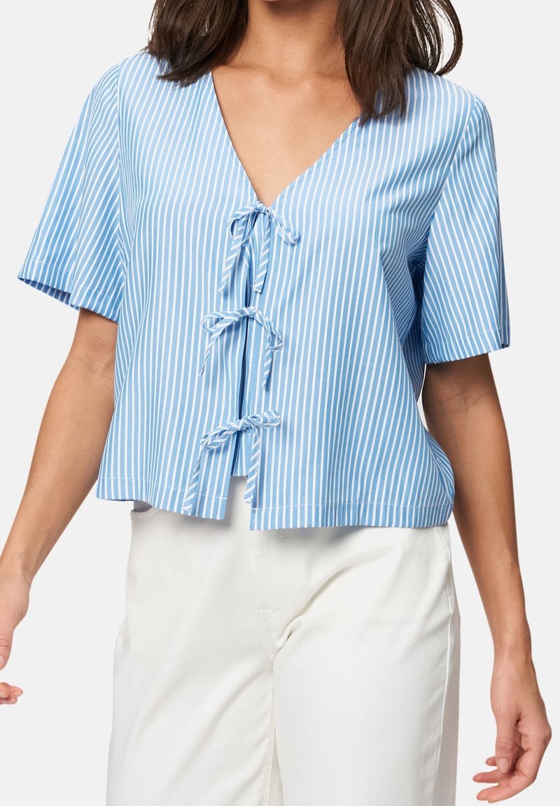 Femme portant un haut à rayures verticales bleues et blanches avec des manches courtes et trois fermetures à nouer à l'avant, associé à un pantalon blanc.