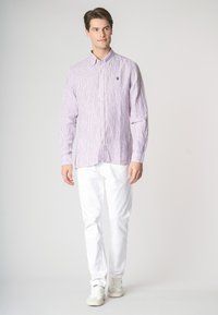 Un giovane uomo con capelli castano chiaro indossa una camicia a righe viola e pantaloni bianchi, in piedi contro uno sfondo semplice, sorridendo con sicurezza.