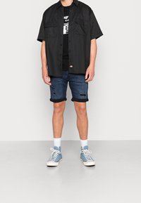 Musta lyhythihainen paita, jossa on kaksi rintataskua, mustan graafisen t-paidan päällä, yhdistettynä sinisiin kulutettuihin farkkushortseihin ja sinisiin lenkkareihin.