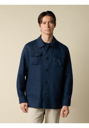 Mann trägt eine dunkelblaue Jacke mit Knöpfen und Brusttaschen, steht vor einem schlichten beigen Hintergrund.