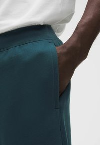 Pantalones de chándal en color verde azulado que presentan una cintura acanalada, bolsillos laterales y una textura suave. Primer plano del material con una mano colocada en el bolsillo.