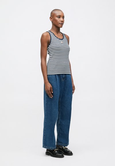 Stāv sieviete ar kailu galvu, ģērbusies melnbaltā svītrainā topā, brīvās zilās džinsās un melnās mokasīnās pret bezkrāsaina gaismas fona.