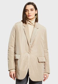 Beige prošívaný blejzr se strukturovaným povrchem, klopami s výstřihem do "V", dvěma předními kapsami a jedním knoflíkem, nošený přes světlý rolák.