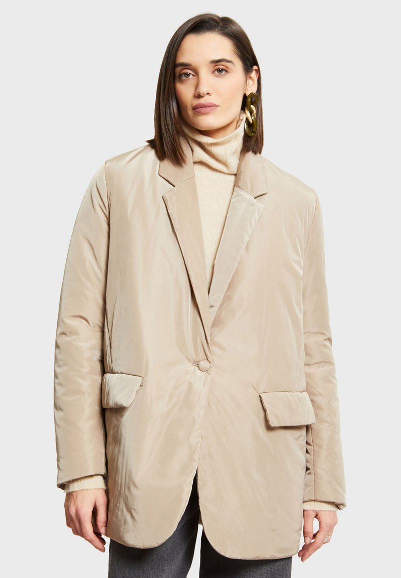 Beige prošívaný blejzr se strukturovaným povrchem, klopami s výstřihem do "V", dvěma předními kapsami a jedním knoflíkem, nošený přes světlý rolák.