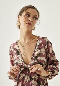 Vestido con estampado floral en malva, con un escote en V profundo y mangas abullonadas. Presenta un detalle de nudo en la cintura. Acentos con collar de cadena dorada.