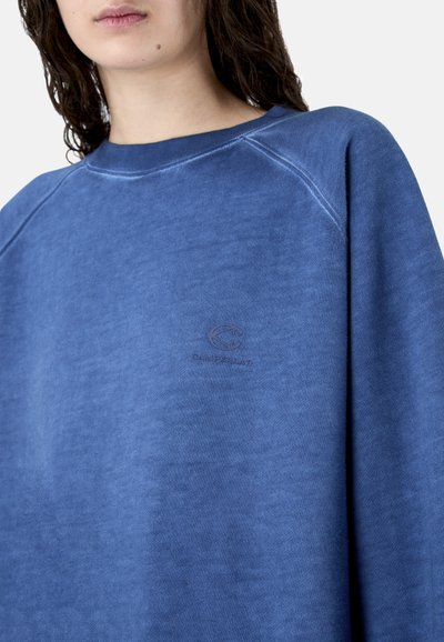 Gros plan sur une personne portant un sweat-shirt bleu avec un petit logo brodé sur la poitrine, montrant les détails de l'encolure et de l'épaule.