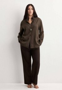 Vrouw met lang donker haar, een loszittend olijfgroen overhemd, donkere wijde broek, sandalen en een hangerketting, staand met handen in de zakken.