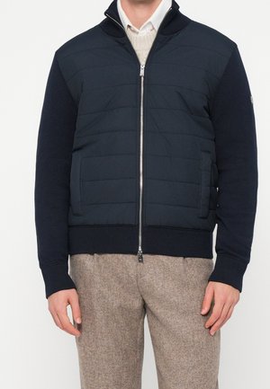 Man draagt een zwarte doorgestikte jas met ritssluiting over een wit overhemd en beige gestructureerde broek, staand met ontspannen armen.