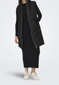 Doudoune noire avec col montant, ornée de coutures diagonales et d'une fermeture éclair, portée par-dessus une robe noire, associée à des sneakers blanches.