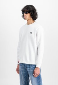 Mann med krøllete hår som har på seg svarte solbriller, hvit sweatshirt med logo og blå jeans, stående mot en ensfarget hvit bakgrunn.