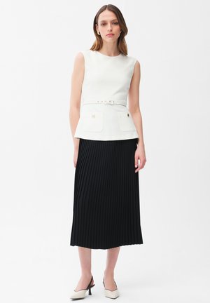 Femme portant un haut blanc sans manches avec ceinture et deux poches avant, et une jupe midi plissée noire, debout devant un fond blanc.