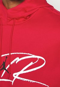 Haut à capuche rouge avec un cordon de serrage, arborant un grand design en forme de swoosh blanc et un logo Jumpman noir. Texture lisse avec une coupe décontractée.