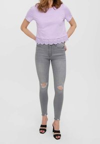 Žena v levandulovém topu s krátkým rukávem a vlnkovaným lemem, šedých roztrhaných skinny džínách a černých sandálech na podpatku, stojící se založenýma rukama za zády.