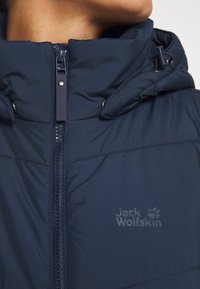 Navy blue puffer jacket with high collar, zip closure, and logo detail. Features smooth texture and reinforced stitching at seams.