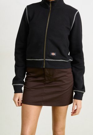 Schwarze Zip-up Sweatjacke mit hellgrauen Nähten, cropped Design und Logo-Patch. Kombiniert mit einem braunen Minirock.