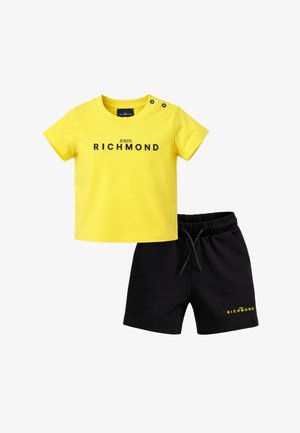 T-shirt gialla a maniche corte con scritta "John Richmond" e bottoni neri sulla spalla, abbinata a pantaloncini neri con coulisse e piccola scritta "Richmond".