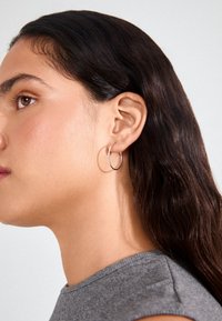 Zilveren ringoorbel te zien aan het oor van een model, met een gepolijste afwerking en circulair ontwerp, dat haar donkere, golvende haar en grijze top aanvult.