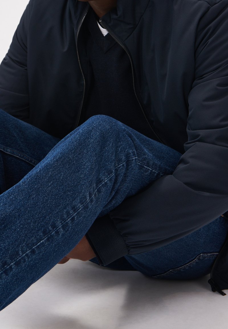 Giubbotto puffer blu navy con chiusura a zip, indossato sopra un maglione nero e abbinato a jeans in denim blu scuro, caratterizzato da una texture liscia e polsini a costine.