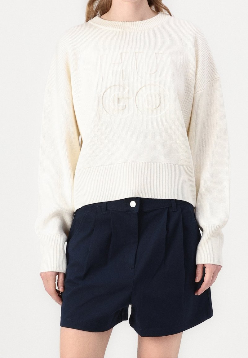 Femme portant un pull côtelé écru avec lettrage en relief « HUGO » et un short taille haute bleu marine avec fermeture boutonnée.
