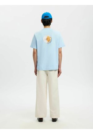 Személy világoskék ingben, melynek hátán taco minta látható, krémszínű bőszárú nadrágban, fekete cipőben és kék sapkában, háttal állva.