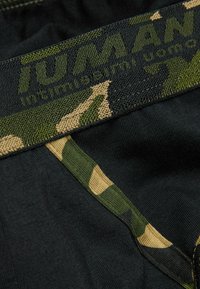 Tejido negro con una cinturilla elástica de patrón de camuflaje. Presenta el logo en verde en la cinturilla. Textura suave, diseño ajustado y bordes sin costuras.
