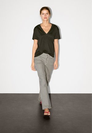 Schwarzes V-Ausschnitt-T-Shirt kombiniert mit gestreiften, weiten Hosen. Das Outfit umfasst rosa Sandalen und hat einen neutralen Hintergrund.