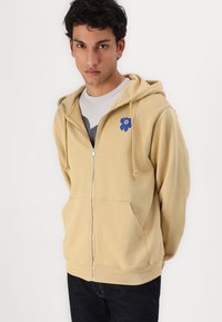 Lys beige zip-up hettegenser med snøring, frontlommer og en blå blomsterlogo på venstre bryst. Myk, glatt tekstur.