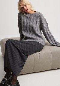 Gray cable knit sweater with a loose fit, paired with a black pinstriped skirt. The seating is a light beige, soft-textured, upholstered design.