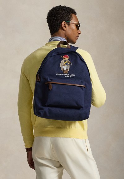 Bărbat purtând un pulover galben și ochelari de soare, cu un rucsac bleumarin cu broderie Polo Bear și accente cu fermoar din piele maro.