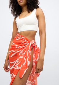 Πορτοκαλί διάφανη φούστα με άσπρα αφηρημένα σχέδια, δεμένη στη μέση, τοποθετημένη πάνω από ένα λευκό εφαρμοστό τοπ. Απαλό, ελαφρύ ύφασμα.