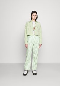 Casaco cropped verde claro com fecho de botão e dois bolsos no peito, combinado com calças verdes com padrão de ondas. Modelo usa sapatos de plataforma pretos.