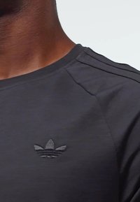 Tmavošedé športové tričko s okrúhlym výstrihom, s textúrovaným čiernym vyšitým logom na hrudi a jemnými detailmi na ramenných švoch.