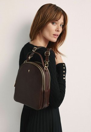 Kobieta o jasnobrązowych włosach, ubrana w czarny sweter, niesie na ramieniu ciemnobrązowy skórzany plecak z złotymi zamkami i klamrami.