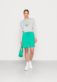 Sudadera gris con estampado gráfico, combinada con pantalones cortos verdes brillantes y zapatillas blancas. El modelo sostiene un bolso verde a juego.