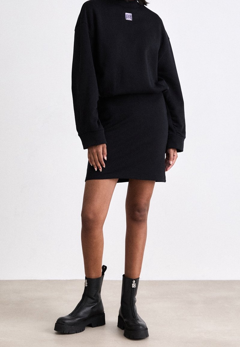 Femme portant un haut noir à manches longues et une jupe assortie, avec des bottes de cheville zippées noires et épaisses, se tenant sur un sol uni.