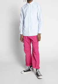 Chemise boutonnée bleu clair à manches longues, associée à un pantalon rose vif et des baskets Converse noires. Comprend une poche poitrine et des poignets retroussés.